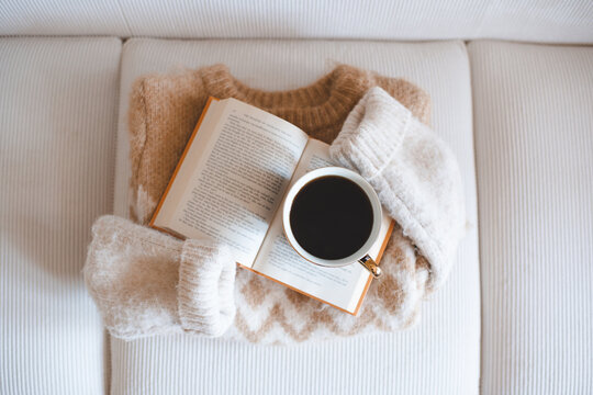 Mug of black coffee on open paper book with folded knitted sweater on couch in living room. Winter cozy atmosphere.