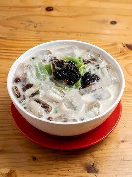 Refreshing Traditional Indonesian Sweet Ice Dessert with Cendol Jelly, Grass Jelly, Coconut Milk, and Ice Cubes in a White Bowl on a Wooden Table