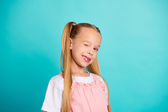 Smiling blonde girl winking with missing tooth in pink dress against blue background, playful child portrait showing joy and confidence