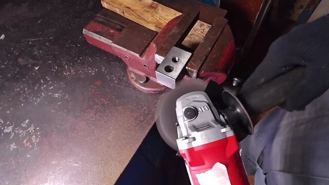 Angle grinder shapes metal beside vise during workshop prep. Gloved hands control red power tool over rusted plate, and grinding action throws bright sparks at end during surface preparation.