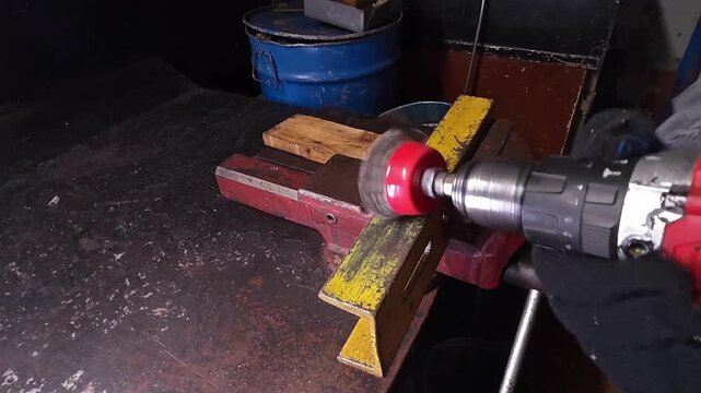 Gloved worker drills metal with handheld tool in workshop vise. Spinning attachment meets clamped stock on bench, showing focused tool use and controlled shaping in dim industrial workspace.