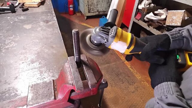 Yellow power grinder shapes steel in workshop repair. Gloved hands control rotating disc near bench vise, and metal part stays secured in busy workshop with worn surfaces and tools.