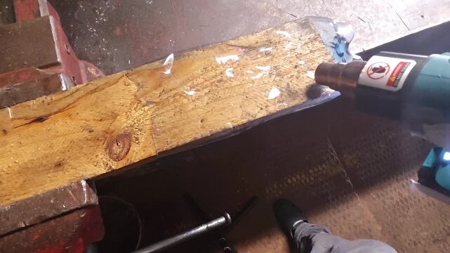 Electric stapler fastens wood on workbench, pressing its heated tip into board during practical repair task. Hands guide tool carefully as wood piece stays clamped in place for secure joining.