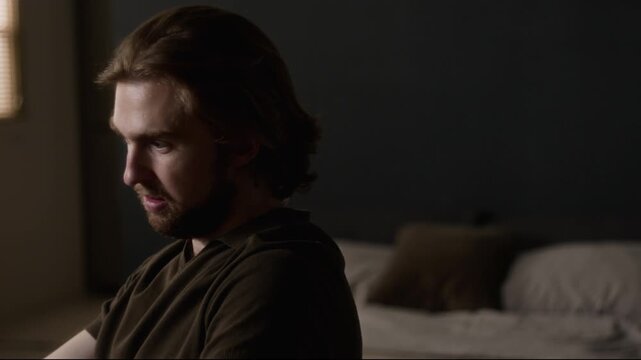 Young adult Caucasian man sitting at home, thinking deeply and clasping hands while typing on laptop with contemplative expression. For mental health awareness, burnout campaigns, wellness advertising