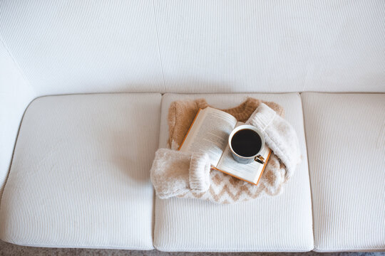Mug of black coffee on open paper book with folded knitted sweater on couch in living room. Winter cozy atmosphere.