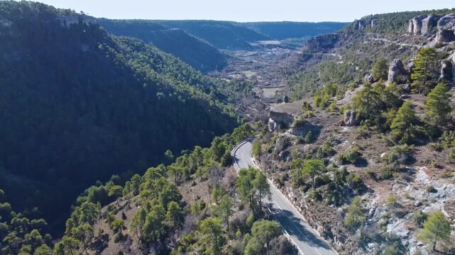 A winding asphalt road curves along a steep rocky canyon cliff edge and dense pine forest (Pinus). Perched at the top of the precipice stands a small ancient stone watchtower overlooking a deep valley