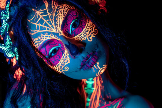 Close-up portrait of woman with glowing neon UV skull face paint, magenta cyan eye designs, golden spiral accents, lace-like cheek lines, and butterfly highlights in dark studio lighting