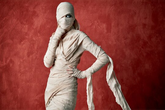 Full length portrait of woman wrapped in textured mummy bandages with one eye visible through narrow facial gap against deep red painted wall in dramatic side light confident standing pose