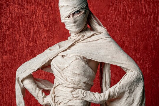 Full body portrait of person wrapped head to toe in textured bandages, posing against deep red painted wall with dramatic side lighting, confident hands on hips studio