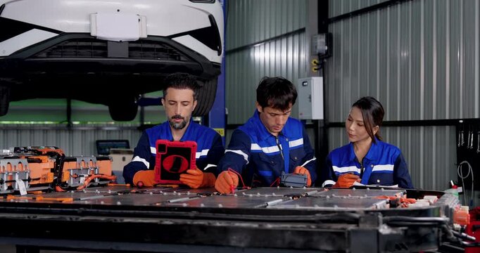 Three EV technicians inspect exposed battery pack beneath raised electric vehicle, using diagnostic tablet, meter and notes while coordinating battery testing in modern automotive service workshop.