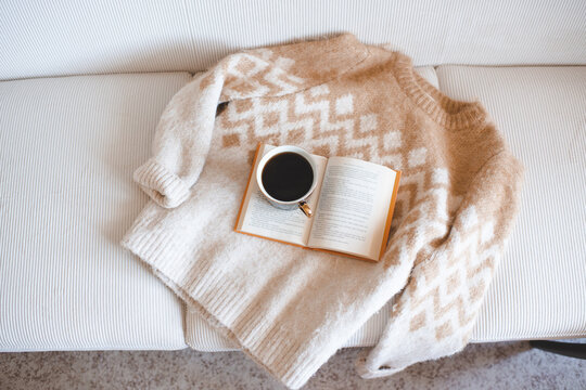 Mug of black coffee on open paper book with folded knitted sweater on couch in living room. Winter cozy atmosphere.