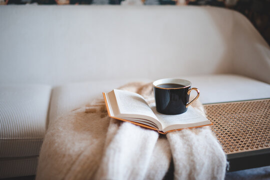 Cup of fresh black coffee staying on open paper book with knitted sweater on rattan table over white couch in living room. Cozy hygge atmosphere.