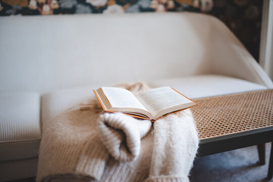 Open paper book on knitted sweater on rattan table over white couch in living room close up. Cozy evening atmosphere. Winter season.