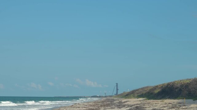 A sandy beach stretches along blue ocean waves under a clear sky near a coastal spaceport. The distant launch tower, dunes, shoreline, and calm daylight show a scenic aerospace landscape.