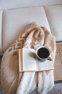 Cup of fresh black coffee staying on open paper book with knitted sweater on rattan table over white couch in living room. Cozy hygge atmosphere.