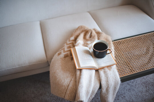 Cup of fresh black coffee staying on open paper book with knitted sweater on rattan table over white couch in living room. Cozy hygge atmosphere.