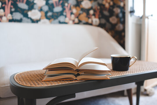 Cup of black tea with stack of open paper books on rattan table in living room close up. Night time.