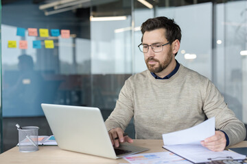 A Focused Businessman Working On A Laptop And Reviewing Important Documents At His Desk In A Modern...
