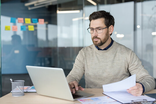 A Focused Businessman Working On A Laptop And Reviewing Important Documents At His Desk In A Modern Corporate Office. The Professional Manager Is Analyzing Financial Data And Planning Strategy While.