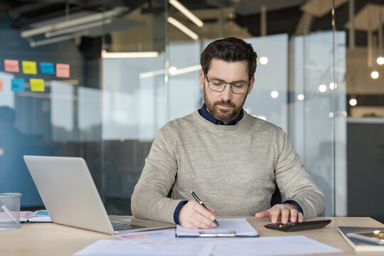 A Focused Businessman Working On A Laptop And Writing Documents For Financial Analysis At A Desk In A Corporate Office. The Professional Manager Is Carefully Reviewing Paperwork And Using.