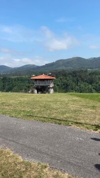 Family walks toward traditional Horreo in Asturias, Spain. Blue sky above rolling green hills. Children laugh as parents guide them along gravel path. Ideal for travel, cultural