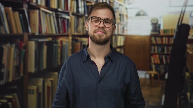 Man with hand on chest waving amid bookshelves in a building library aisle, wearing glasses and a navy shirt, smiling; gratitude warmth.