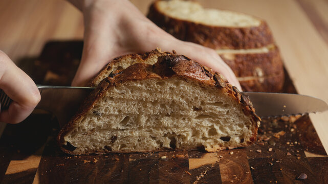 Close-up of hands slicing freshly baked artisan sourdough bread with a knife on a wooden cutting board. Homemade crusty open crumb loaf for breakfast, bakery promos, and healthy food concepts.