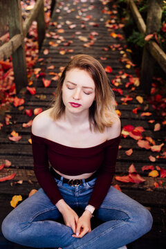 Young woman with closed eyes sitting on wooden pathway covered with autumn leaves, enjoying nature, reflecting in peaceful atmosphere. Feeling harmony, reunion with nature. Relaxing, mental recovery