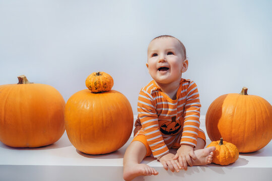 Halloween background with little surprised Caucasian baby in orange costume with lantern jack sitting among pumpkins on white background. Autumn fall harvest festival concept. Copy space
