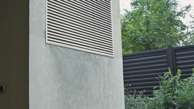 An air vent mounted on a building wall, transitioning through various angles. The setting features foliage and a fence, showcasing the blend of indoor and outdoor spaces