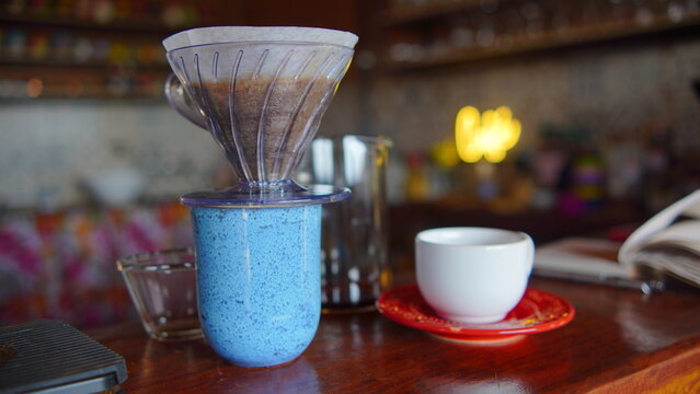 Freshly brewed pour over coffee dripping through a glass filter into a blue ceramic mug in a cozy cafe setting