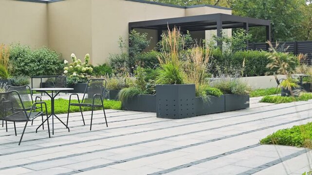 The garden features a wooden deck surrounded by various plants and flowers in large planters. Tables and chairs are positioned on the paving stones, creating a usable outdoor space