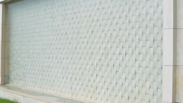 A glass wall with a textured pattern with a waterfall illuminated by natural light. The wall is part of a modern architectural setting where greenery is visible at the base