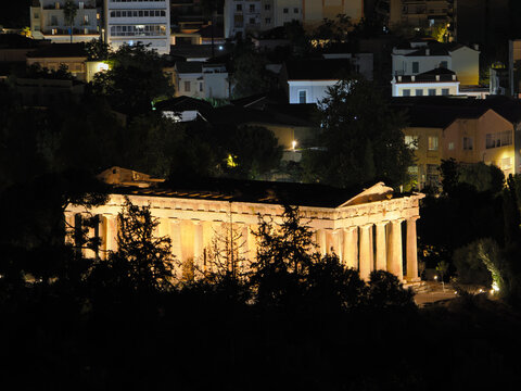 Classical Greek Hephaestus temple lit up at night with residential buildings visible in the dark background, Athens, Greece