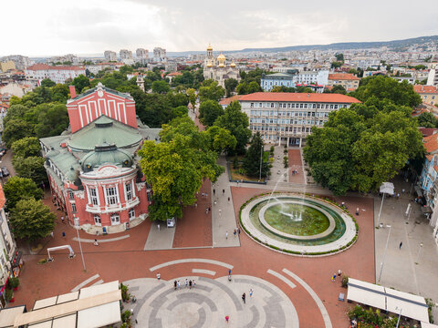 Aerial view of Opera Varna with its red facade and green roof, overlooking the central square with a circular fountain, surrounded by trees and urban architecture in Varna, Varna, Bulgaria.