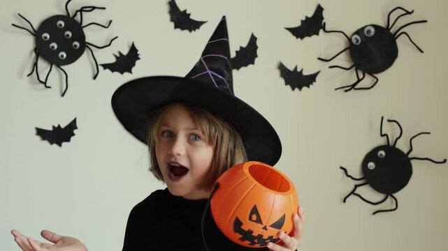 Video portrait happy excited face girl trick or treat Halloween surprise kid wearing dress witch hat costume party Halloween. Toddler holding pumpkin jack lantern basket candies trick or treating