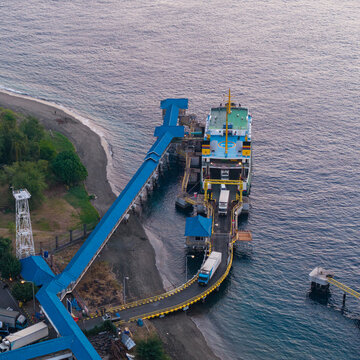 Aerial view of Gilimanuk Port with a ferry docked, trucks on the ramp, and a long blue pedestrian walkway extending along the coastline in Kecamatan Melaya, Bali, Indonesia.