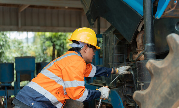 Asian male agricultural engineer in safety helmet using air nozzle gun to clean tractor engine. Skilled mechanic carrying out routine machinery maintenance in outdoor garage.
