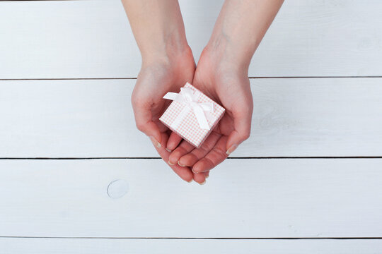 Female hand holding pink gift in hands on a white wooden table. View from the top.
