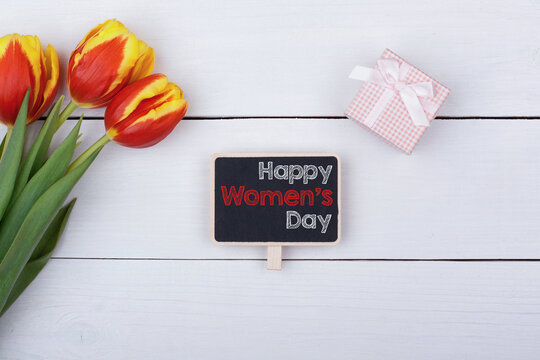 Blackboard with Happy Women's Day, bouquet of tulips and a gift with a bow on the table.