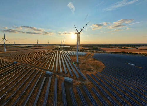 Aerial view of Chelveston Renewable Energy solar and wind farm at sunset with golden hour light illuminating rows of solar panels and turbines in Chelveston, England, United Kingdom.