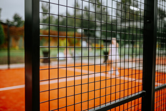 Padel court with orange surface and black fencing, featuring blurred players engaged in a match, surrounded by green trees and additional courts visible in the background