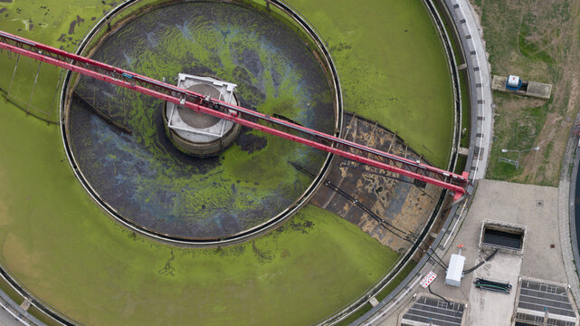 Aerial view of a circular sedimentation tank at a wastewater treatment plant with green algae and a red bridge structure in Cologne, North Rhine-Westphalia, Germany.