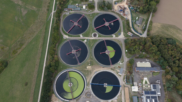 Aerial view of a wastewater treatment plant with circular sedimentation tanks and industrial buildings surrounded by green fields and trees in Cologne, North Rhine-Westphalia, Germany.
