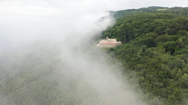 Aerial view of Mare de deu de la salut Hermitage, dramatically perched on a rocky cliff above dense forests, overlooking the beautiful mountain landscapes of Girona, Catalonia. 