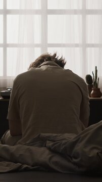Vertical video of young adult Caucasian man waking up, rubbing neck, getting out of bed, rear view. Clip suits mental wellbeing ads, fatigue stories, sleep healthcare campaigns