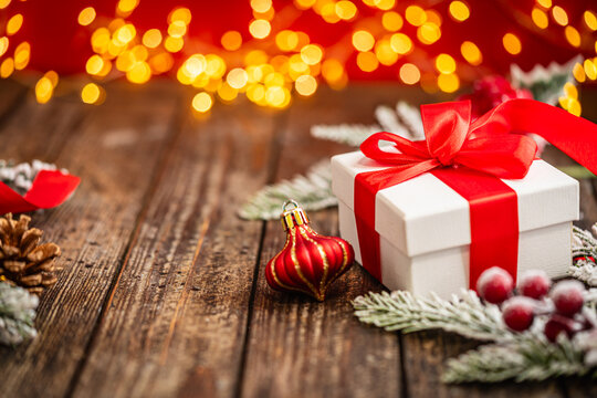A Christmas background featuring a white gift box tied with red ribbon beside a pinecone, red ornaments, and frosted berries on rustic wood, lit by warm twinkling lights above with copy space.