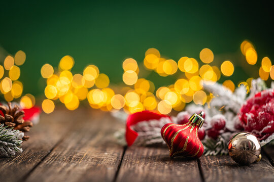 A Christmas background featuring a red plaid bauble with a snowflake and gold ribbon, red ornaments, and frosted berries on rustic wood, set against green with twinkling lights and copy space.