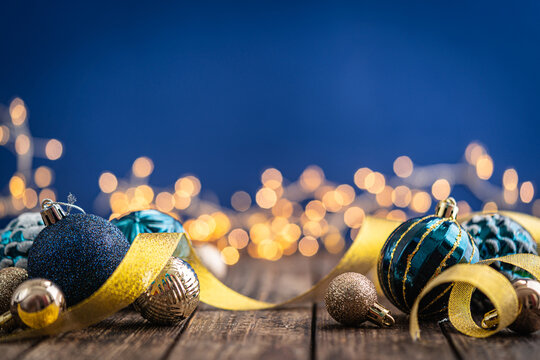 A Christmas background featuring navy and teal ornaments wrapped with gold ribbon on rustic wood, framed symmetrically against a deep blue backdrop with twinkling lights and copy space.
