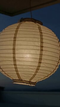 Vertical video: Swaying paper lantern on porch at dusk, breeze causing spin, internal LED glowing
